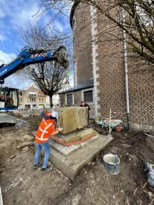 Project Oorlogsmonument Zonhoven, het onderste deel van het monument wordt met een blauwe kraan terug geplaatst met hulp van een man in oranje veiligheidsjas met helm en een werkman.