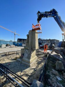 Op de markt van Zonhoven staat het gedeelte waar het monument afgezet met een lage en zwarte omheining. De arm van de blauwe kraan is bezig met het bovenste gedeelte van het monument.