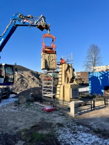 Het bovenstel deel van het monument wordt terug geplaatst door de blauwe kraan. Er ligt wat sneeuw op de voorgrond. Het is mooi weer, de zon staat laag en er is heel wat schaduw.
