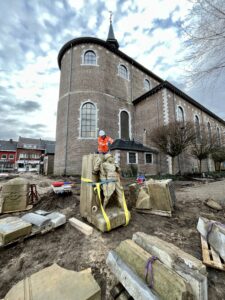 Een bewolkte lucht met de kerk van zonhoven. Het oorlogsmonument staat vooraan op de foto omringd door de resterende delen van het monument en bouwmateriaal. Een man in oranje veligheidsvest en helm staat achter het monument.